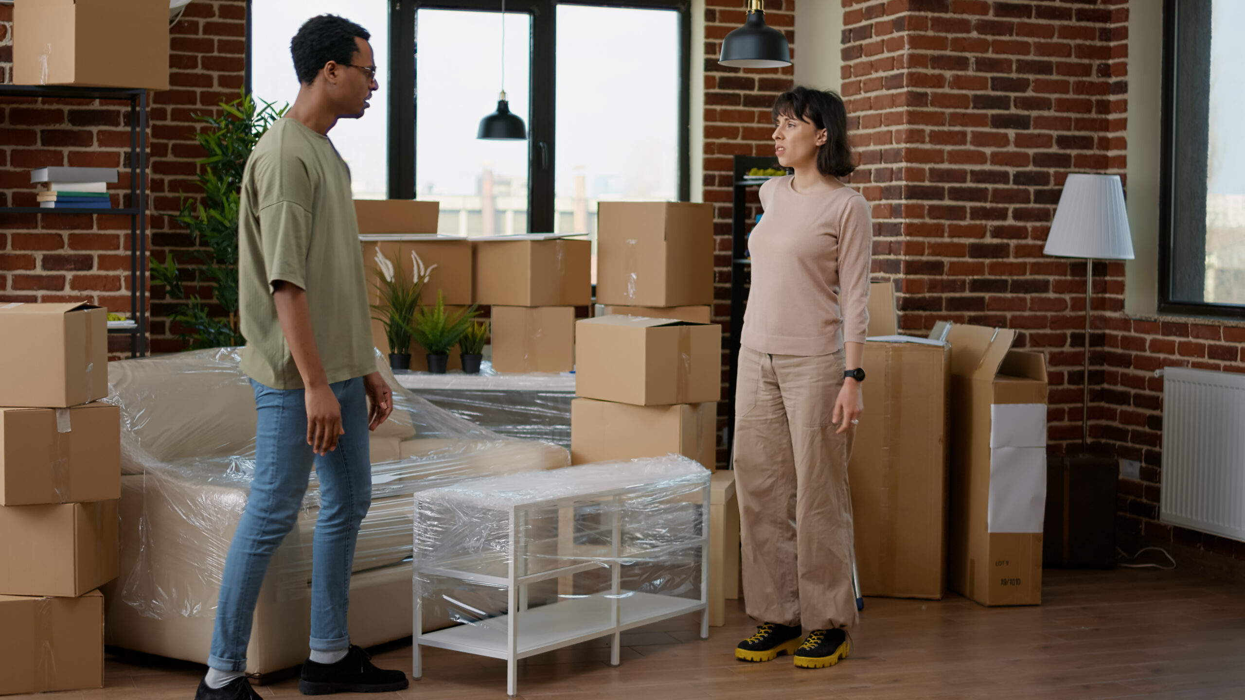 Two movers dressed in orange overalls, one pushing a cart with cardboard boxes and the other carrying a rolled rug, while loading items into a moving truck outdoors.