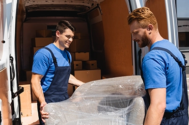Two professional movers in blue uniforms carrying a piece of furniture wrapped in clear plastic near a moving van.