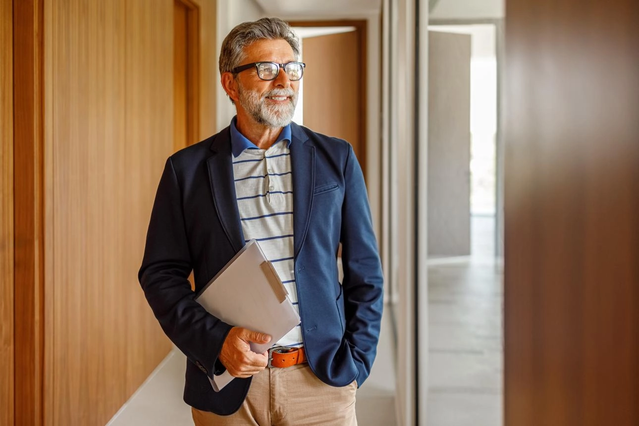 Middle-aged apartment owner wearing a blazer and glasses holding documents while standing in a modern apartment building hallway.