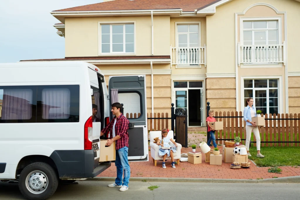 A family unloading boxes from a moving van in front of their new home, with a child and woman carrying boxes while a man unloads from the van, preparing to settle in.