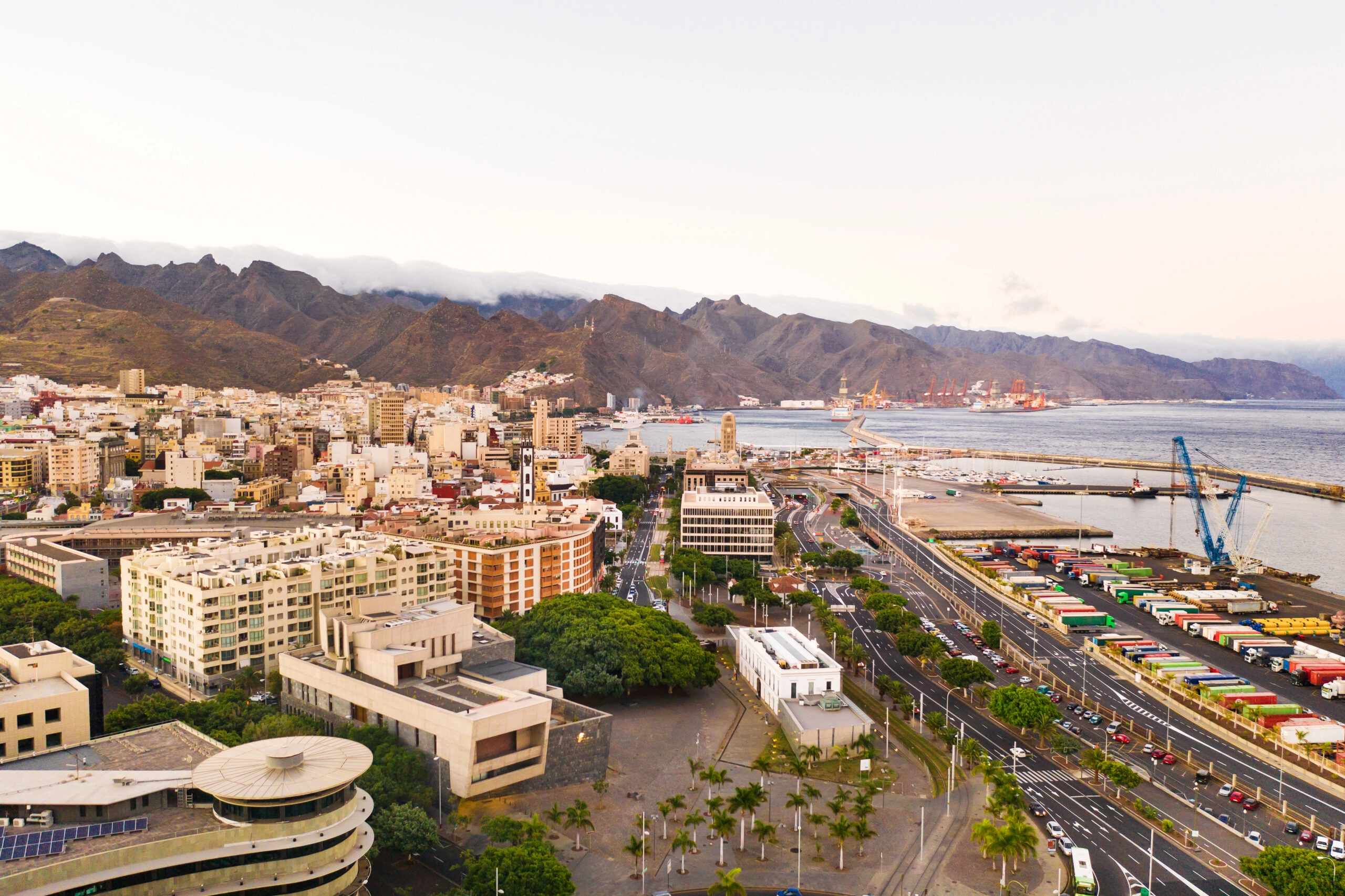 A wide landscape view of a village with green palm trees, a large stone fort, and dry mountains in the background.